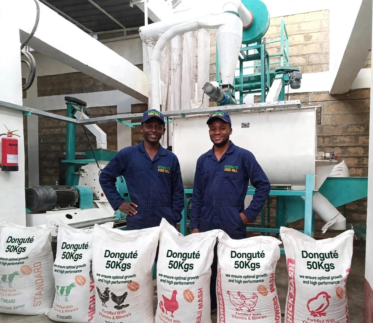 Mr Anutele Samuel and Mr Kilofenyuy Bruno and Madame Neba Christelle Gwin in the Donguté feed mill, in branded overalls behind stacked 50 kg feed sacks